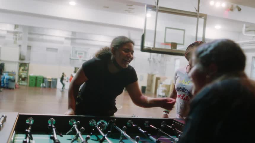 Teenage girls play with foosball table as youths play basketball in the background