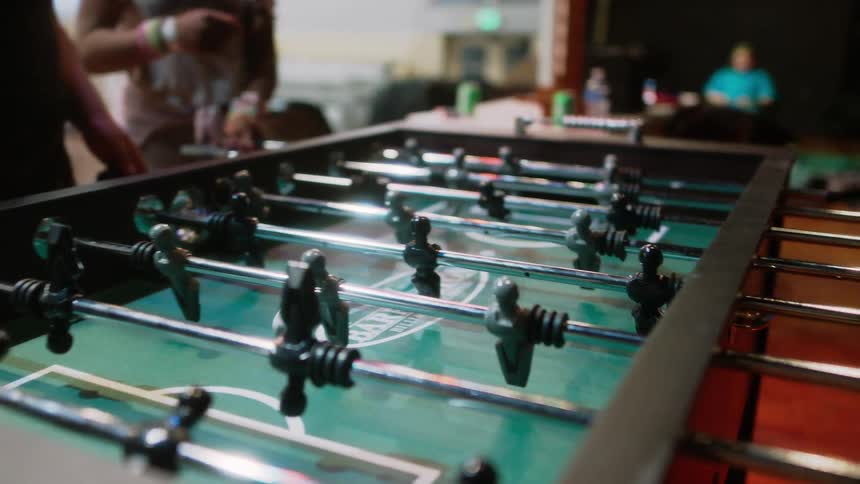 A group of friends play foosball inside of a recreation area with other people around