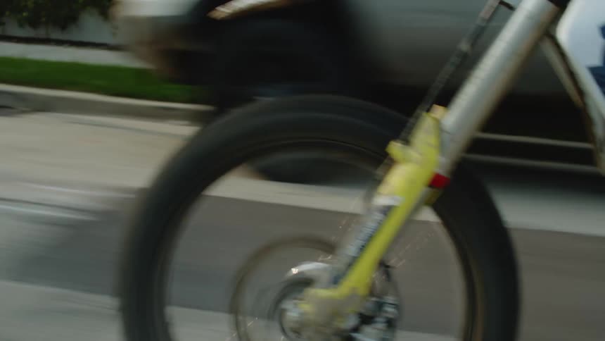 Motorcyclists with a white helmet stands up on the bike while speeding on a street past parked vehicles