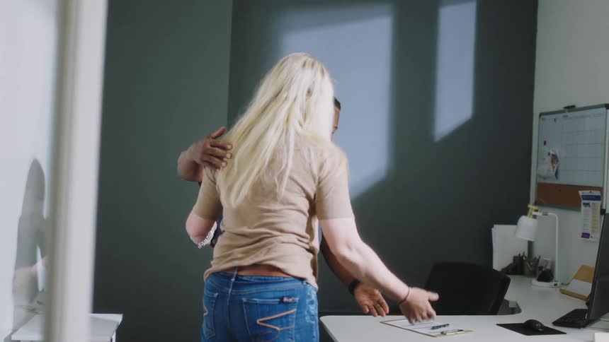 Blonde woman approaches a black male and they hug then they sit down at a desk with a computer on it