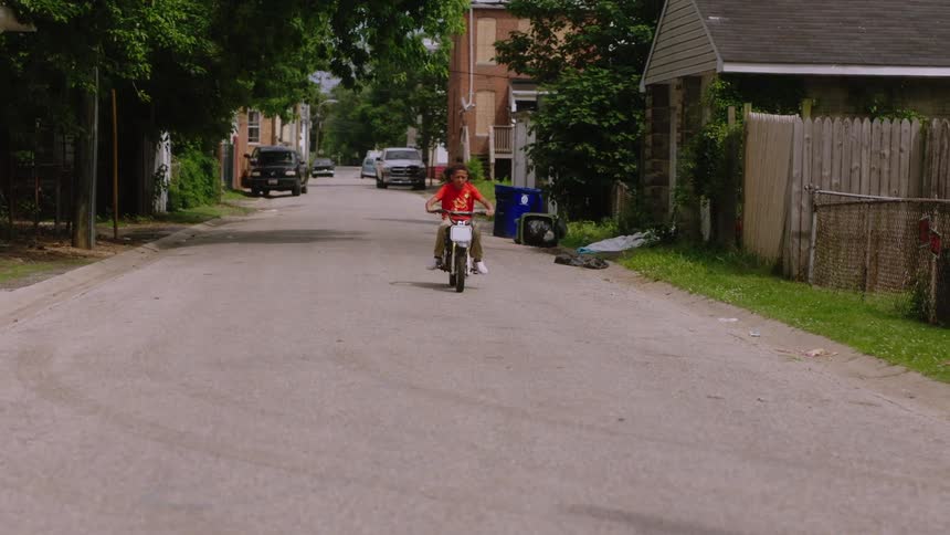 Teenaged boy rides a small motorcycle past some fencing on a side street