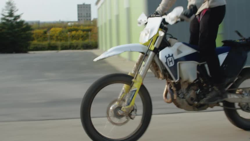 A rider pops a wheelie on his dirt bike as he races past commercial buildings and parking lots