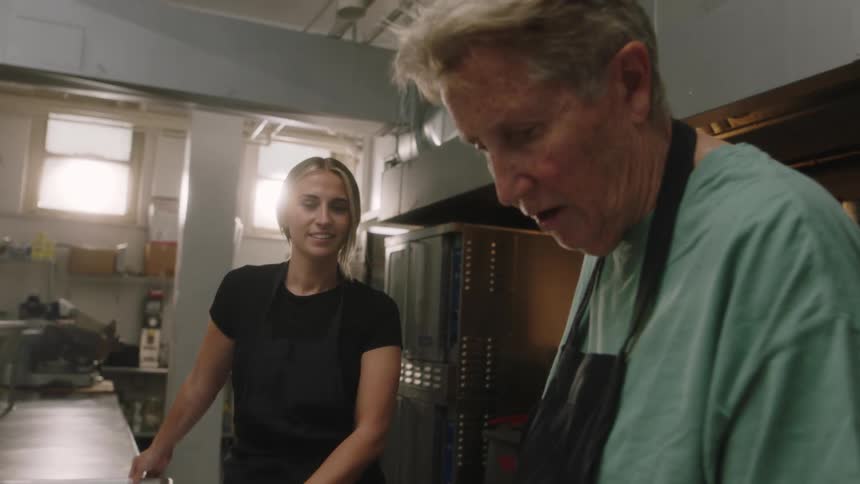 Elderly man uses a knife while preparing food while a woman carries a tray with food on it into the restaurant kitchen
