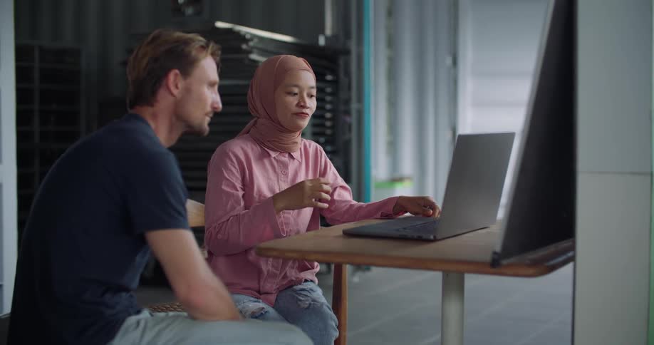 A woman wearing a hijab is sitting in front of a laptop computer talking to man sitting next to her