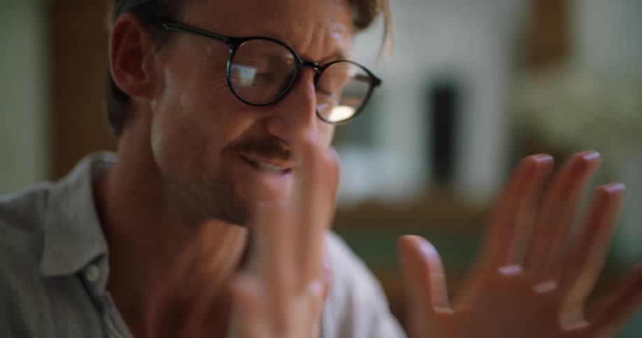 A man is sitting indoors waving to somebody and he puts on his glasses as he looks emotional