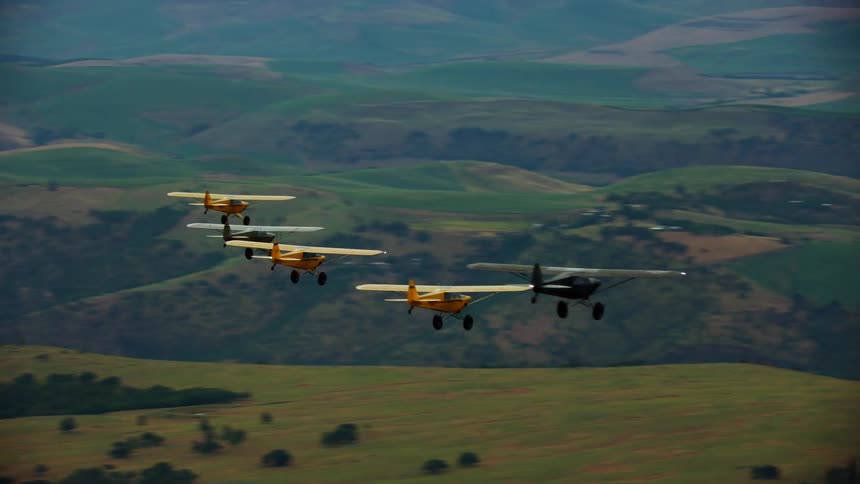 A group of small backcountry airplanes fly in formation over a hilly countryside during the early morning
