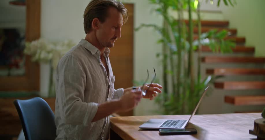 A man is sitting at a table typing on a laptop computer and he stops to take off his glasses and rubs his eyes