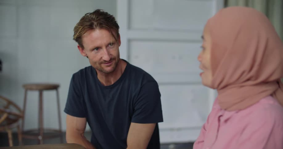 White man and middle eastern woman sitting together at the table and holding a conversation while using a laptop