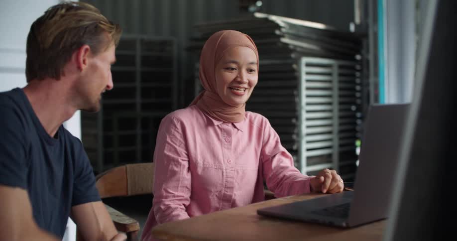 A woman wearing a hijab is sitting in front of a laptop computer talking to man sitting next to her as they point at the screen