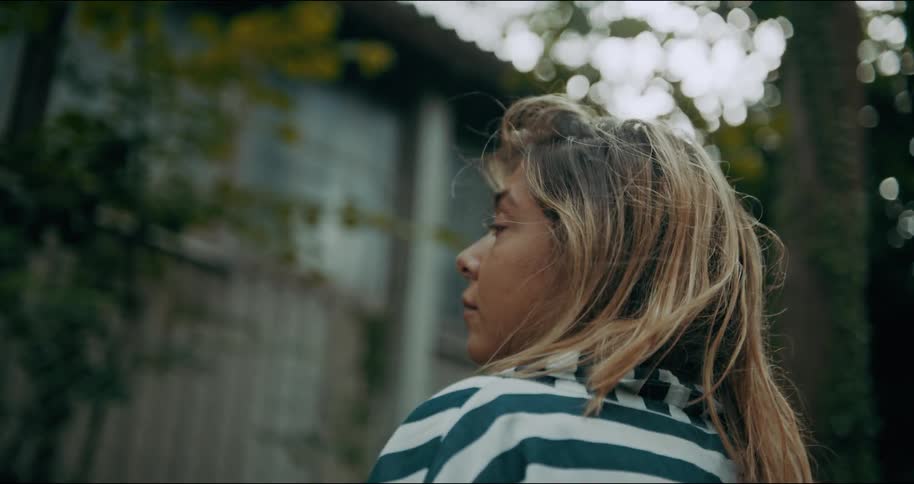 A hispanic woman with blonde hair wearing gold hoop earings and a navy striped shirt stands looking up at windows on a building with the breeze blowing through her hair