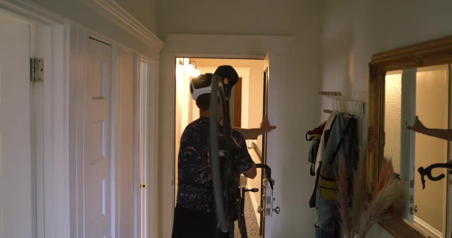 Man wearing shorts and a bicycle helmet carries a bike on his shoulder and leaves a room