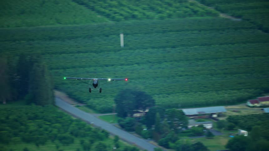 A single engine backcountry airplane with flashing lights on its wings flies above crop fields and vineyards