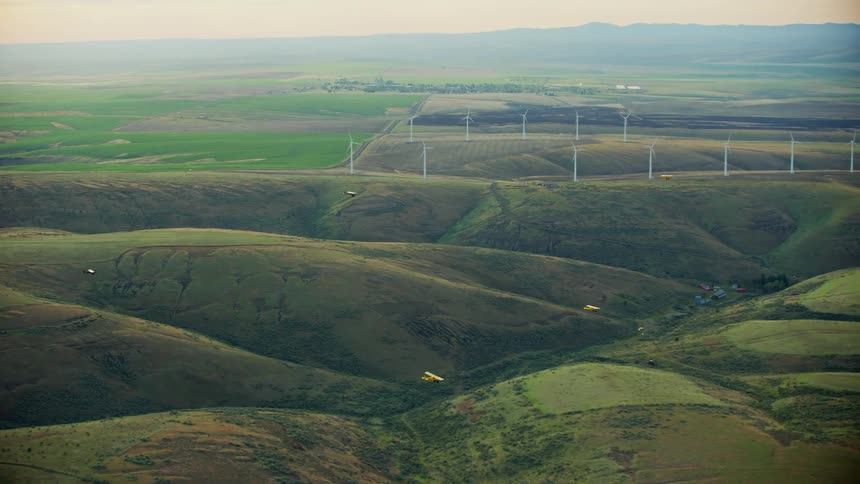 A group of small backcountry airplanes fly past a wind farm in a hilly countryside during the day