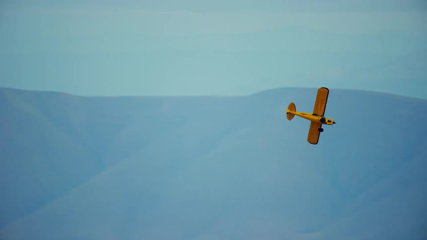 A small yellow backcountry airplane flies with another plane near mountains in a hazy sky