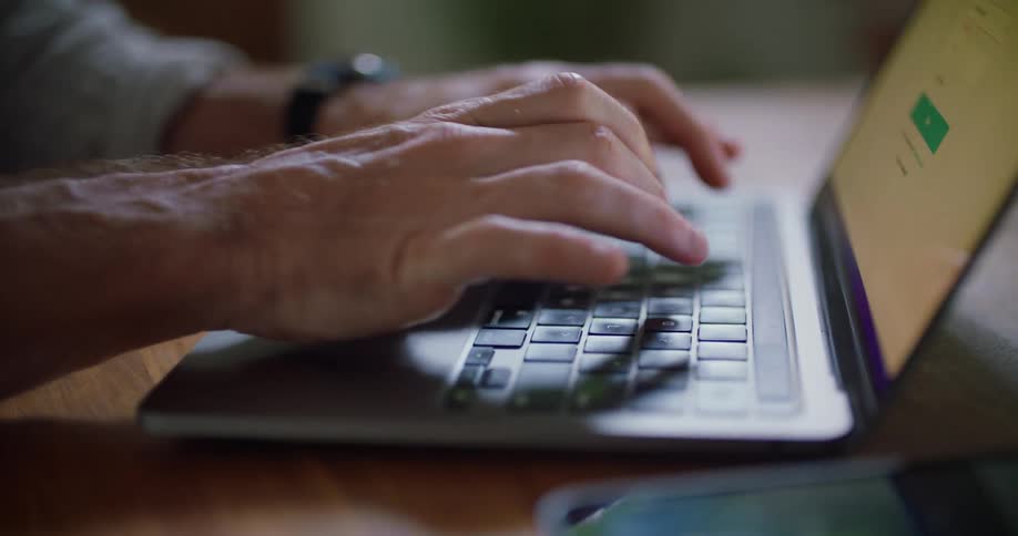 An older white man wearing a black watch is typing the keys on a laptop