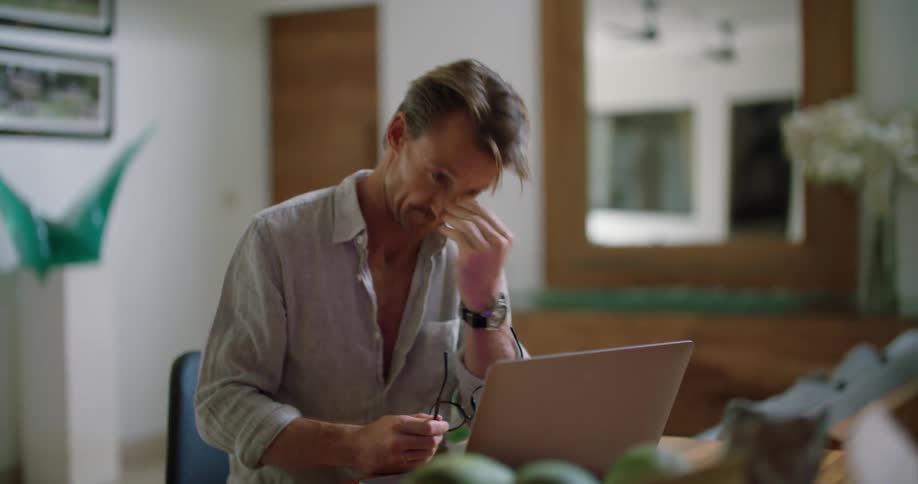 A man is sitting at a table typing on a laptop computer keyboard and he stops to take off his glasses and rubs his eyes then puts his glasses back on and continues