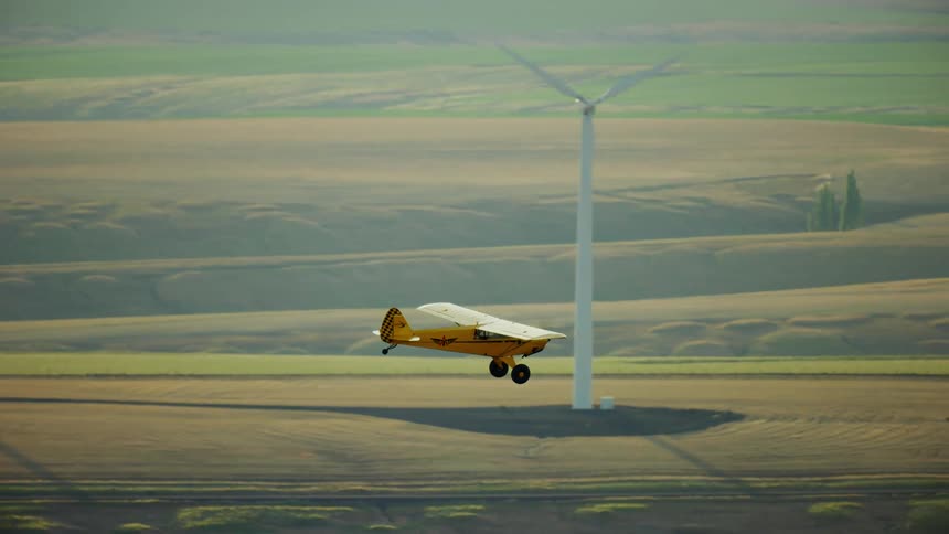 A small yellow backcountry airplane flies low near spinning wind turbines on a wind farm