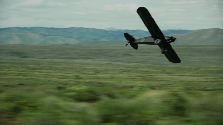 A small backcountry airplane is flying low near the grass covered ground and there are mountains in the distance