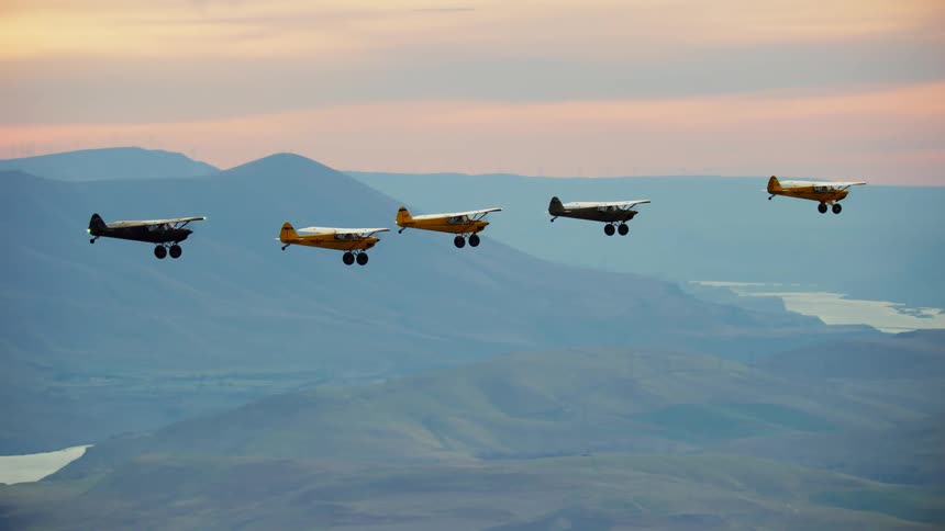 Backcountry airplanes fly in formation through a red tinted sky and over river in misty valley surrounded by mountains