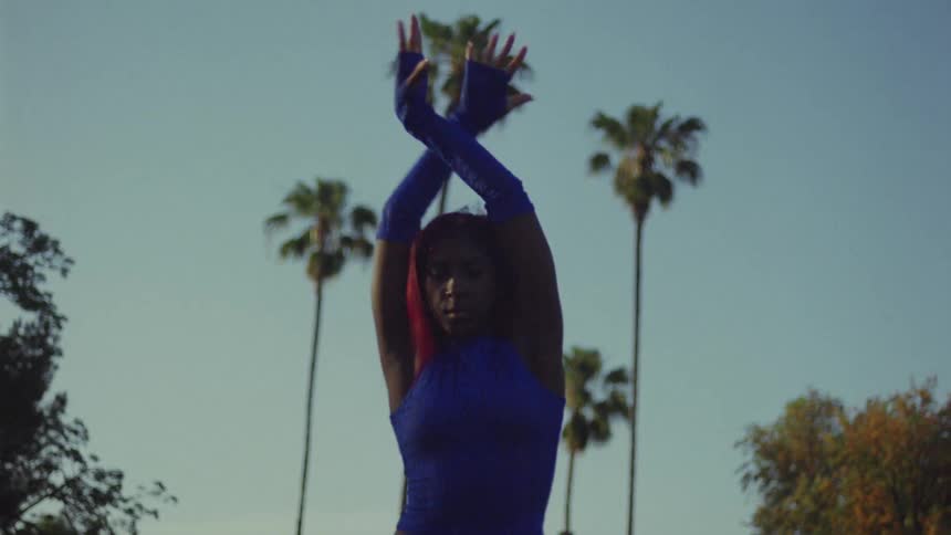 Woman wearing blue gloves dancing outdoors with palm trees in the background