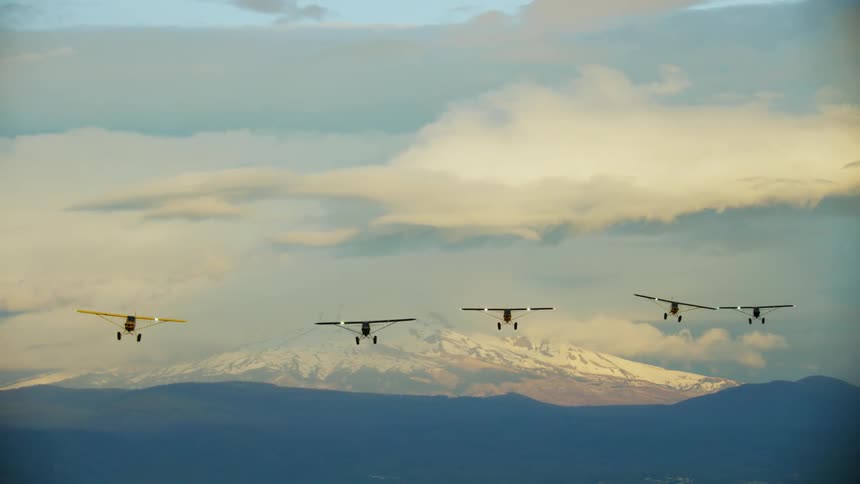 A group of single engine airplanes fly over a grassland toward a snow capped mountain in the distance during the day