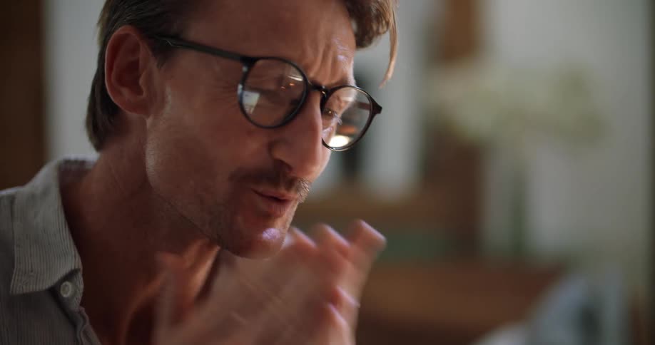Man smiles and blows kisses at laptop screen reflected in his glasses before rubbing his eyes tiredly
