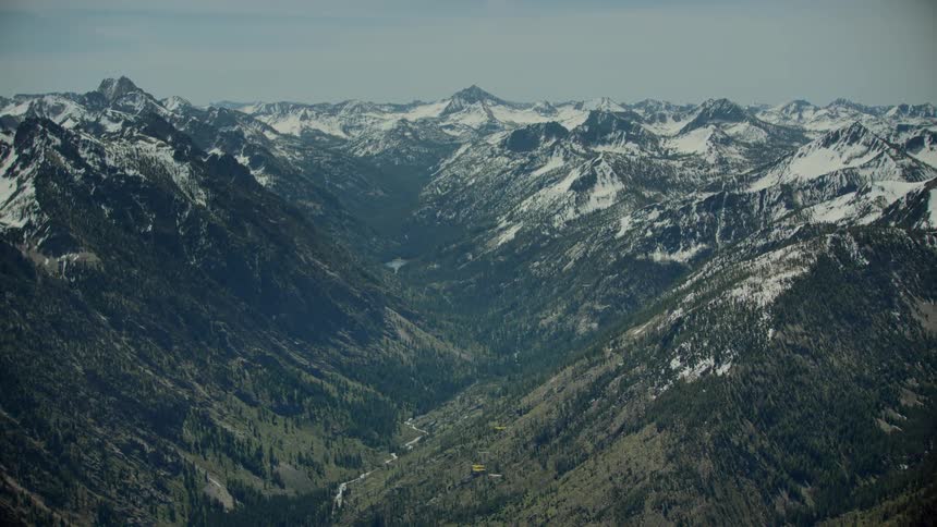 A river runs through the valley between snow covered forested mountains and there are mountains in the distance