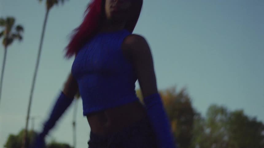 Black woman wearing long blue gloves and a matching blue top moves her arms as she dances in a field near the palm trees