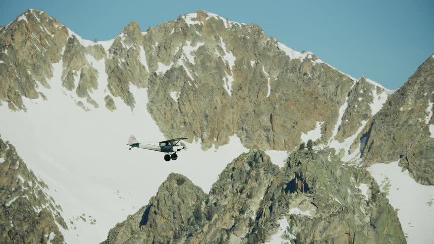 A small backcountry airplane flies near snow covered mountains with peaks high in a clear sky in the Sawtooth mountains near Stanley Idaho
