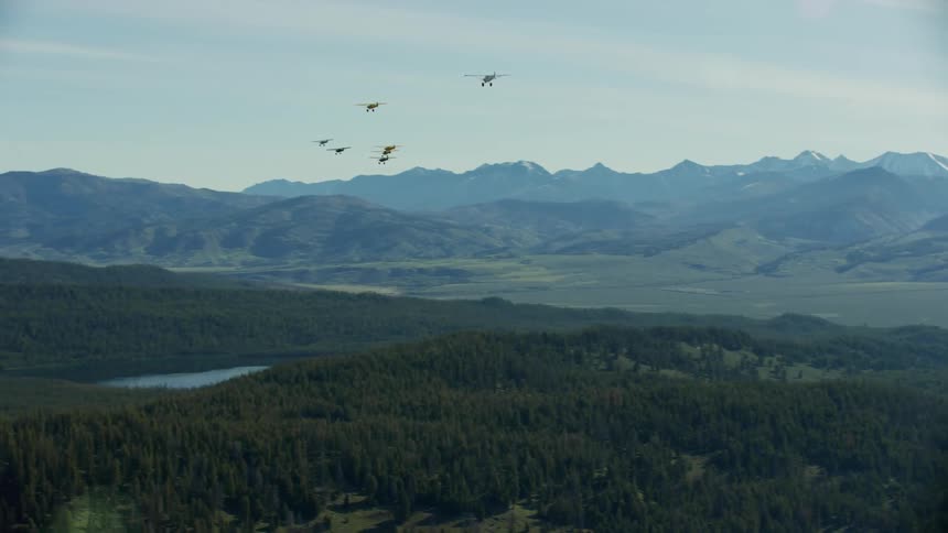 Several backcountry airplanes with fixed landing gear fly over water and forest in valley surrounded by mountains on misty day