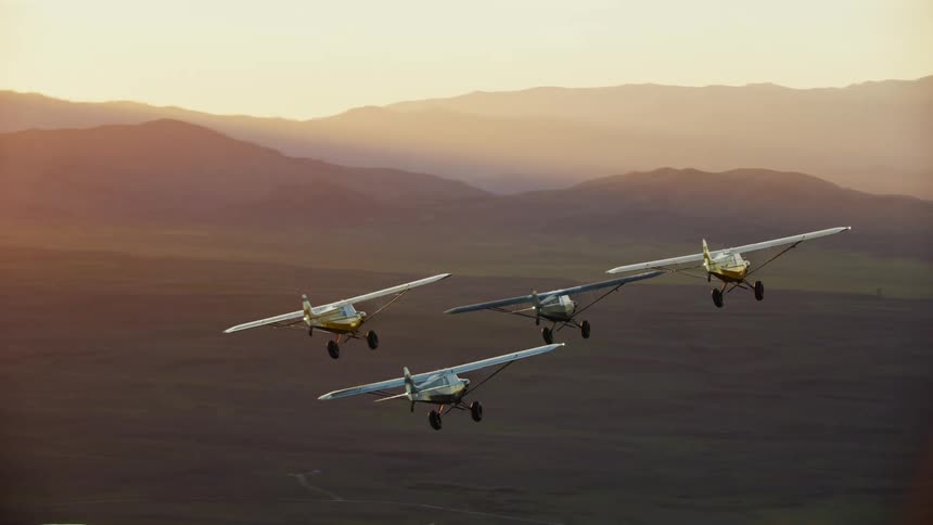 A group of small backcountry airplanes fly in formation over the countryside during the day