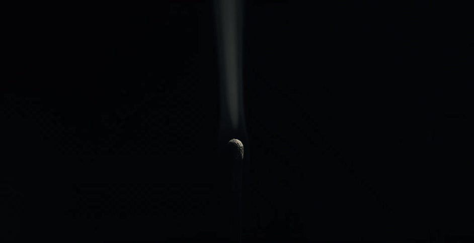 Smoke puffs and rises from an extinguished match head against black
