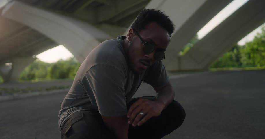 A man kneeling under a bridge with one hand on the pavement raises his head to look into the camera