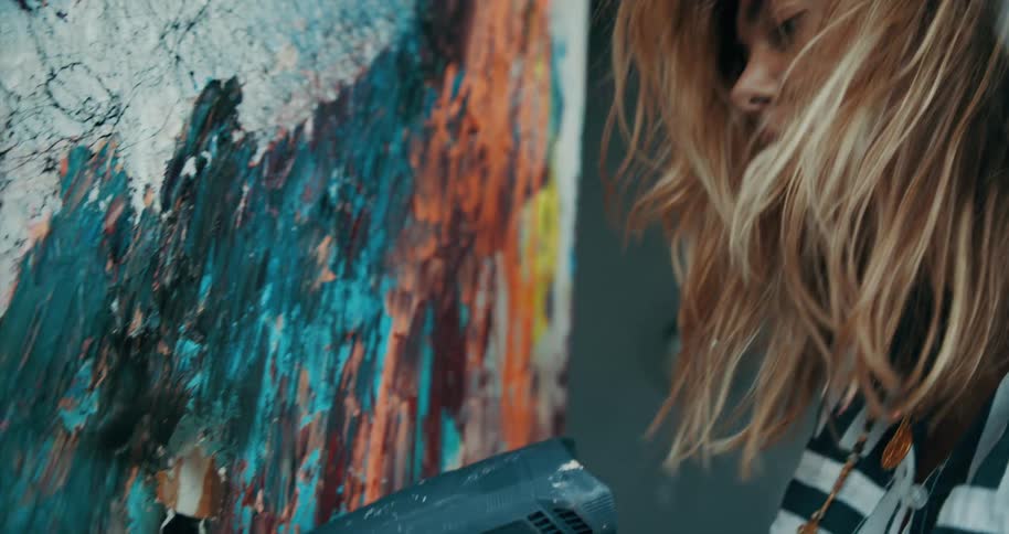 A hispanic woman with blonde hair is holding a heat gun that is heating up colorful paint on a canvas
