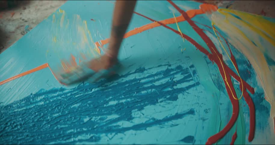 Artist kneeling on the floor of a studio paints with bright colors over canvas using her hands