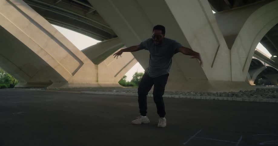 A black man wearing dark colored jeans and shirt with glasses and white sneakers is dancing under a bridge