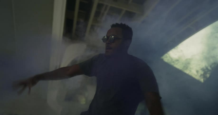 Man wearing sunglasses dances surrounded by blue smoke under a highway