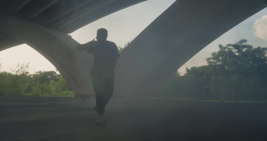 A dark skinned male wearing pants and a tshirt is dancing under a bridge with smoke all around him