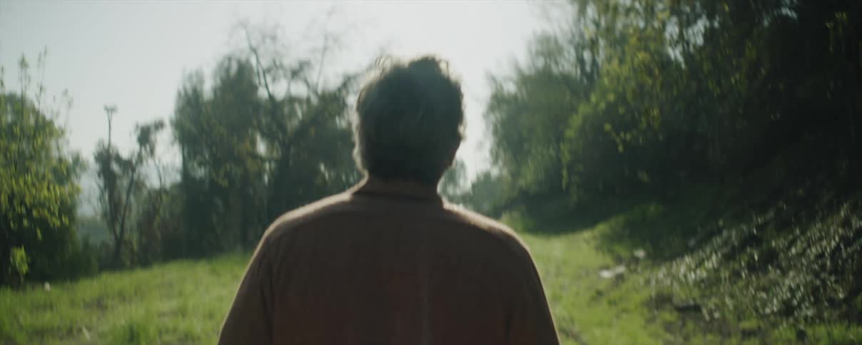 A man is outside walking on a dirt road with lined with trees and the sun is shining brightly