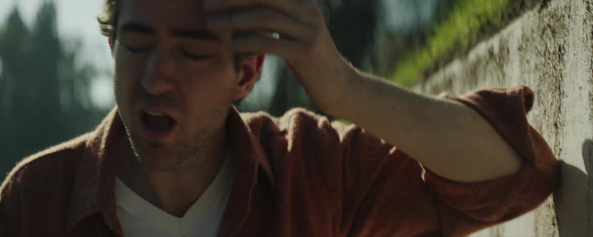 A man is outside walking on a sidewalk near a wall embankment and he stops to lean against the wall and rub his face