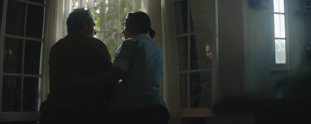 A old man and woman in a blue outfit with brown hair in a ponytailsit next to a window staring at each other sadly and hugging each other