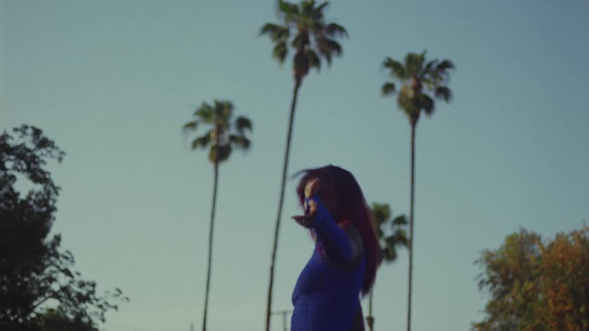 Black woman wearing long blue gloves and a matching blue top dances outdoors near the palm trees