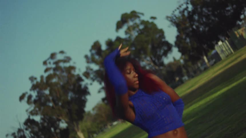 Black woman wearing long blue gloves and a matching blue top moves her arms as she dances in a field on a sunny day