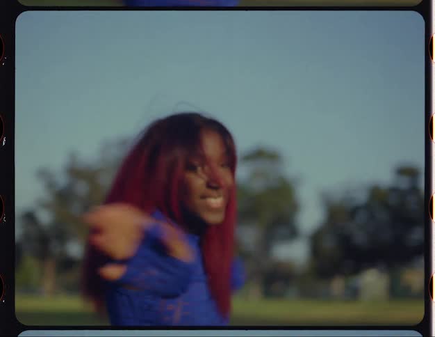 A young woman smiles into the camera while dancing in a field during the day