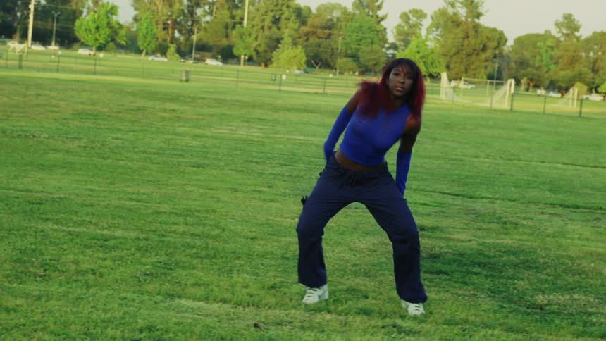 A teenage girl is outside dancing in a field as traffic moves by on the street in the distance