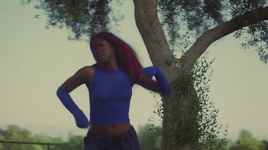 Woman with red hair blue top and sports pants dances on lawn in public park