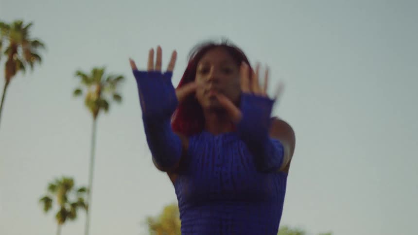 Black woman wearing a blue top and matching blue gloves moves her arms while she dances near the palm trees