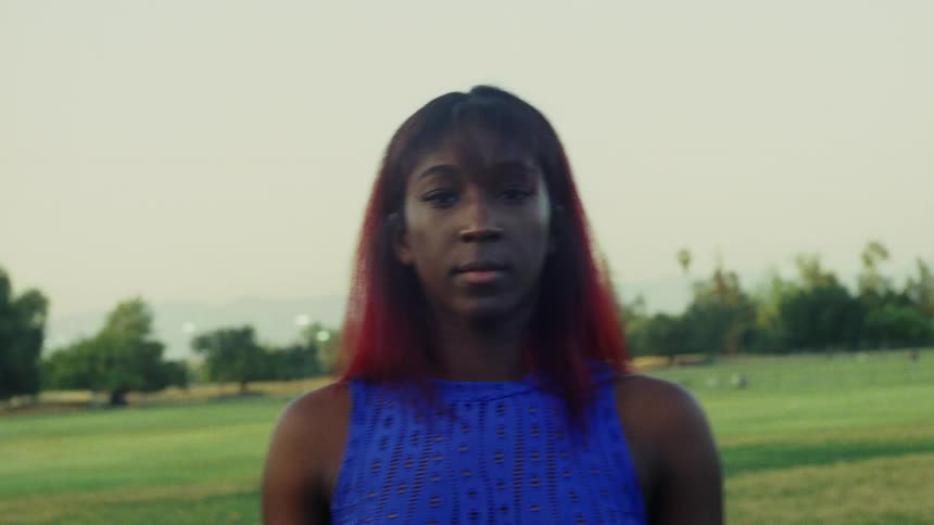 A young woman looking into the camera while standing in a field glances away before returning her gaze to the camera