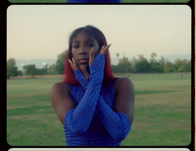 Black woman wearing long blue gloves and a matching blue top moves her arms as she dances while standing in a field