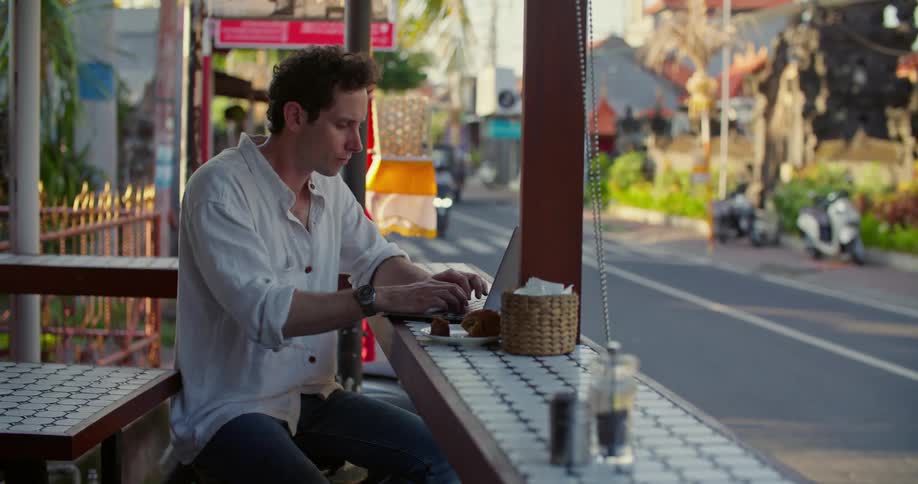 Man sits on restaurant porch typing while traffic passes by on nearby street